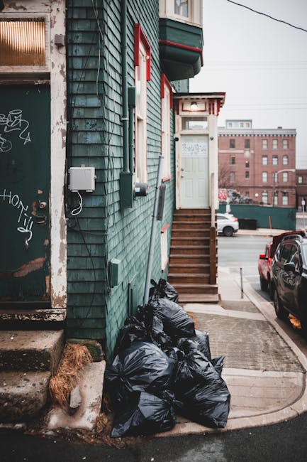 A close-up view of the side of a residential building featuring green wooden cladding with a textured, weathered finish. Several black rubbish bags, filled and tied, are stacked on the pavement near the building's corner, indicating disposal of household waste. The building has a small, white door at the top of a short flight of wooden steps with a handrail, and red trimming around the door and window frames; the steps lead from a narrow sidewalk to an entrance elevated above the street level. To the left, a portion of an adjacent door with graffiti markings and a weathered surface is visible, alongside a small patch of insulation or damaged material at the base of the wall. In the background, across the street, there are low-rise brick buildings with flat roofs and various vehicles, including parked cars. The scene is illuminated by natural daylight, depicting an urban environment prepared for private rubbish collection by rubbishclearancetufnellpark.com, reflecting typical local waste disposal practices for bulky items or overfilled rubbish.