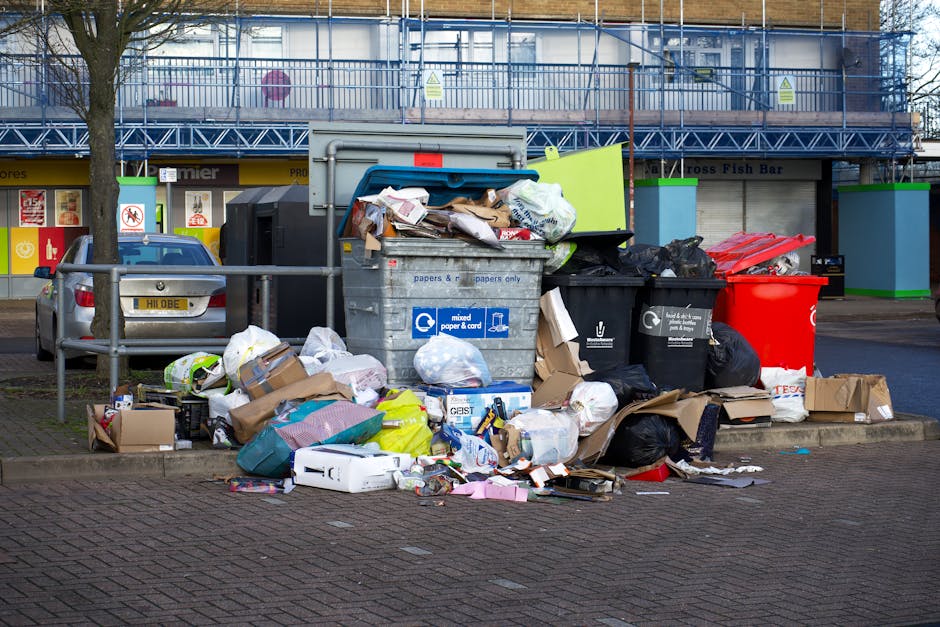 A broad view of a large pile of mixed waste and rubbish bags accumulated around a grey recycling bin on a paved urban street. The waste includes cardboard boxes, plastic bags, paper, and various packaging materials, some spilling onto the surrounding pavement. The recycling bin is partially open with loose paper and packaging protruding from the top, and it displays labels indicating it is for mixed paper and card. Behind the rubbish, there is a parked silver car on the left side and a metal railing separating the waste area from the adjacent sidewalk. In the background, a building with scaffolding and various storefronts suggests an urban commercial area. The scene is illuminated by natural daylight, with a tree visible on the left, and the environment indicates an area where private or independent rubbish removal services, such as those provided by rubbishclearancetufnellpark.com, might be employed for alternative waste collection and on-site clearance.
