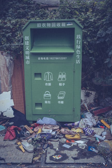 A close-up view of the side of a residential building featuring green wooden cladding with a textured, weathered finish. Several black rubbish bags, filled and tied, are stacked on the pavement near the building's corner, indicating disposal of household waste. The building has a small, white door at the top of a short flight of wooden steps with a handrail, and red trimming around the door and window frames; the steps lead from a narrow sidewalk to an entrance elevated above the street level. To the left, a portion of an adjacent door with graffiti markings and a weathered surface is visible, alongside a small patch of insulation or damaged material at the base of the wall. In the background, across the street, there are low-rise brick buildings with flat roofs and various vehicles, including parked cars. The scene is illuminated by natural daylight, depicting an urban environment prepared for private rubbish collection by rubbishclearancetufnellpark.com, reflecting typical local waste disposal practices for bulky items or overfilled rubbish.