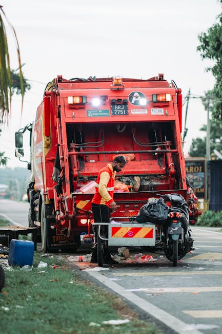 A worker dressed in a yellow and red uniform is operating a red rubbish collection vehicle, likely a garbage truck, on a roadside. The back panel of the truck is open, revealing mechanical components and the interior of the waste collection system. The worker is leaning forward, handling a black plastic bag of waste, which is partially placed in a designated compartment at the rear of the vehicle. The scene shows a paved road with a white parking line, a small curb, and a grassy verge on the left side. In the background, there are trees, utility poles, and a street sign, indicating an urban or suburban setting. The area appears well-lit with natural daylight, suggesting daytime conditions. The presence of waste materials on the ground and the worker’s activity reflects a typical scene of on-site waste collection, possibly part of an alternative or private rubbish removal service. The image emphasizes professional waste management practices and highlights the use of dedicated waste collection equipment, aligning with the context of rubbish removal services such as those offered by rubbishclearancetufnellpark.com for local shops and commercial premises.