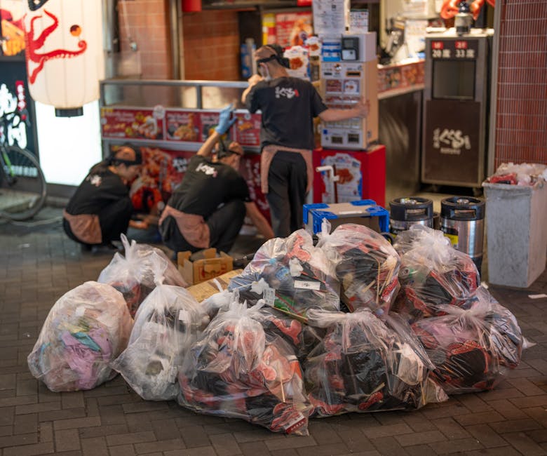 A scene of urban waste collection featuring multiple large transparent plastic bags filled with various types of rubbish, including clothing and packaging, situated on a paved sidewalk. Behind the bags, three workers dressed in dark uniforms with brown aprons and gloves are kneeling and standing near a storefront, which displays bright signage and menu boards. One worker is leaning forward, possibly lifting or arranging waste, while another appears to be managing a piece of equipment or leaning against the counter. Their clothing and the presence of gloves indicate an organized rubbish removal process, likely part of a private collection service such as those offered by rubbishclearancetufnellpark.com. The environment suggests a busy commercial area, with the pavement leading to a shopfront that features a red brick exterior and illuminated signage. The scene is lit by surrounding street and storefront lighting, giving a neutral and professional tone which aligns with an on-site rubbish clearance operation, emphasizing collection and disposal outside a retail or service establishment.