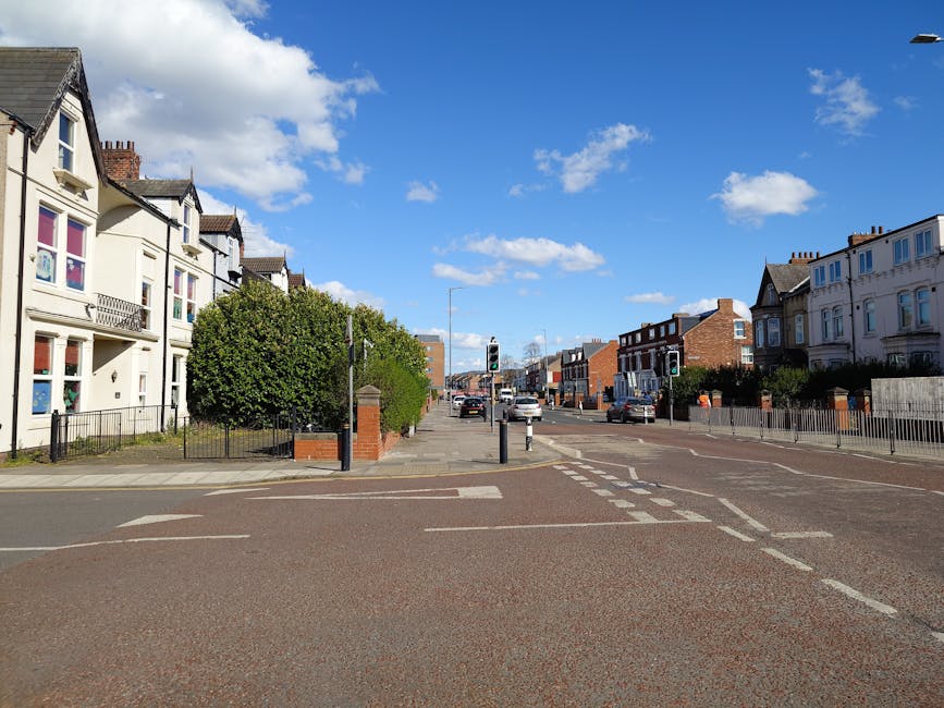 A daytime street scene showing a wide, paved urban intersection with multiple lanes marked by white dashed lines and traffic signals hanging above the crosswalk. On the left side, there is a row of white, Victorian-style terraced houses with pitched roofs, bay windows, and small front gardens enclosed by black metal railings, one of which has a neatly trimmed tree. The right side features a similar row of residential buildings, some with brick facades, and small driveway or garden areas with fences. Several cars, including sedans and hatchbacks, are visible in the distance, either waiting at the traffic lights or driving through the intersection. The sky is bright blue with scattered fluffy white clouds, and the scene is well-lit with natural sunlight, casting minimal shadows. The setting captures a typical neighborhood environment with a mix of residential architecture and active vehicle movement, reflecting an alternative waste handling context that might involve private rubbish collection services near Tufnell Park.