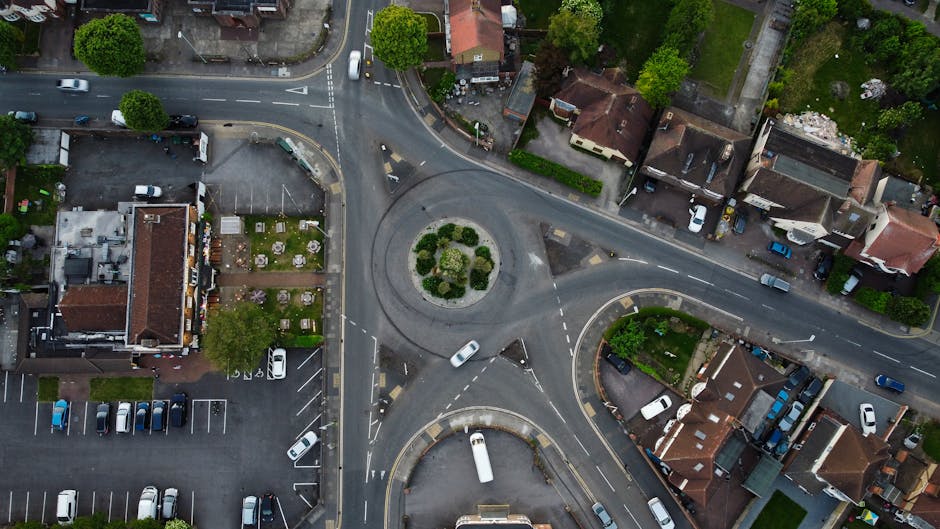 An aerial view of a small roundabout featuring a landscaped central island with neatly trimmed bushes and small trees. Surrounding the roundabout are three converging roads with marked lanes and dashed white lines, showing moderate traffic flow including cars, vans, and small trucks. To the left of the roundabout, there is a large building with a brown tiled roof and an adjacent parking lot filled with parked vehicles, including passenger cars and a few vans. Behind this building, a small green garden area with outdoor furniture and flower beds is visible, characterized by sitting areas and lush greenery. To the right of the roundabout, residential houses with red or brown tiled roofs and small front gardens are lined along the streets, some with parked vehicles on driveways and on the roadside. Trees and shrubs line the residential streets, adding natural greenery to the area. The scene is illuminated by natural daylight, with shadows cast by the trees and buildings, creating a calm urban environment that could support private waste collection or on-site clearance activities in line with professional rubbish removal services like those provided by rubbishclearancetufnellpark.com.