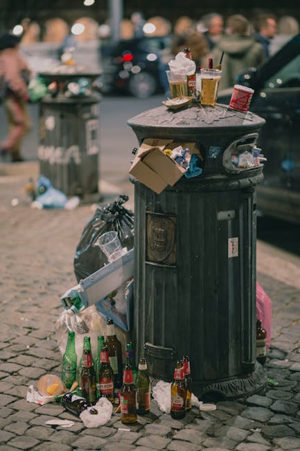 A black, cylindrical public rubbish bin made of cast iron with a domed lid and vertical ridges along its surface, positioned on a cobbled street. Various litter and waste are visibly accumulated around the base, including several glass bottles, some with labels still attached, and crumpled paper or cardboard. Several used paper cups and food containers are placed on top of the bin, with some overflowing and spilling their contents onto the lid. The background features a parked car on the right side of the image, and a blurred view of pedestrians and other urban elements, such as a large trash bin and additional litter, indicating a busy street scene. The lighting suggests daytime with natural light, and the scene reflects urban waste issues that professional rubbish removal services like those offered by [COMPANY_NAME] can address through on-site clearance and private waste handling to promote cleaner public spaces.